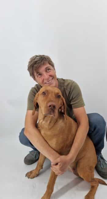 Eine Person mit kurzen Haaren, grünem Hemd und Jeans kniet und umarmt einen großen braunen Hund vor einem schlichten weißen Hintergrund.