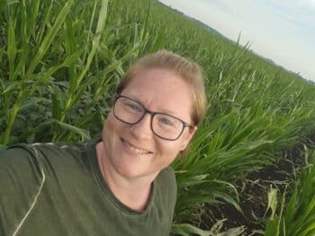 Eine Person mit Brille und grünem Hemd lächelt, während sie ein Selfie in einem grünen Kornfeld unter einem teilweise bewölkten Himmel macht.