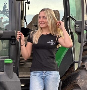 Eine Frau in schwarzem T-Shirt und Jeans steht auf den Stufen eines grünen John-Deere-Traktors, hält ihr Haar und schaut zur Seite.